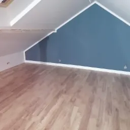 Poddasze po remoncie: drewniana podłoga, pomalowana na biało skośna ściana z drewnianą belką i fragmentem okna dachowego, oraz niebieska ściana szczytowa z białym wykończeniem przy suficie.