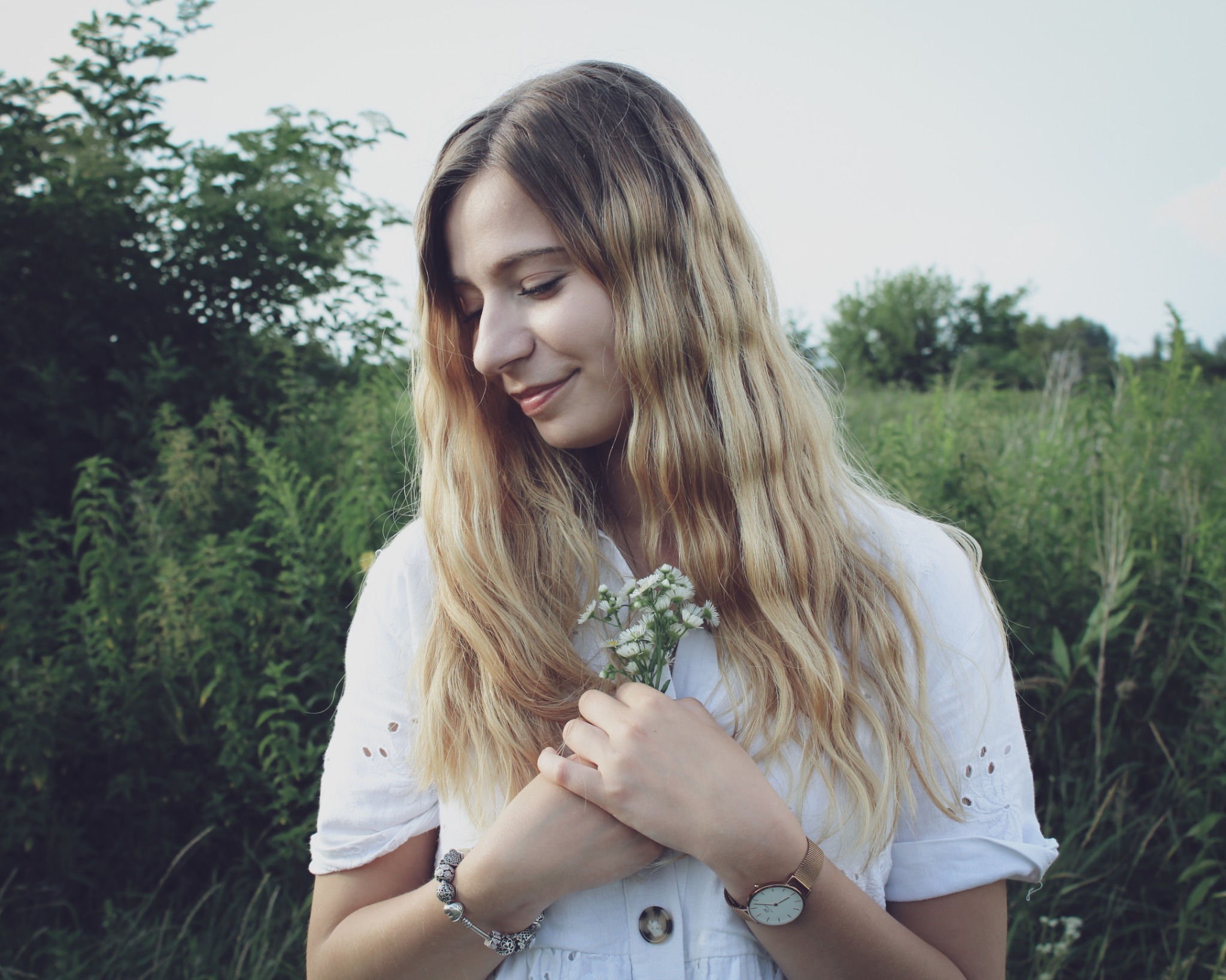 Uśmiechnięta młoda kobieta z blond włosami w białej sukience trzyma bukiecik białych kwiatów na tle zieleni, z delikatnym oświetleniem.