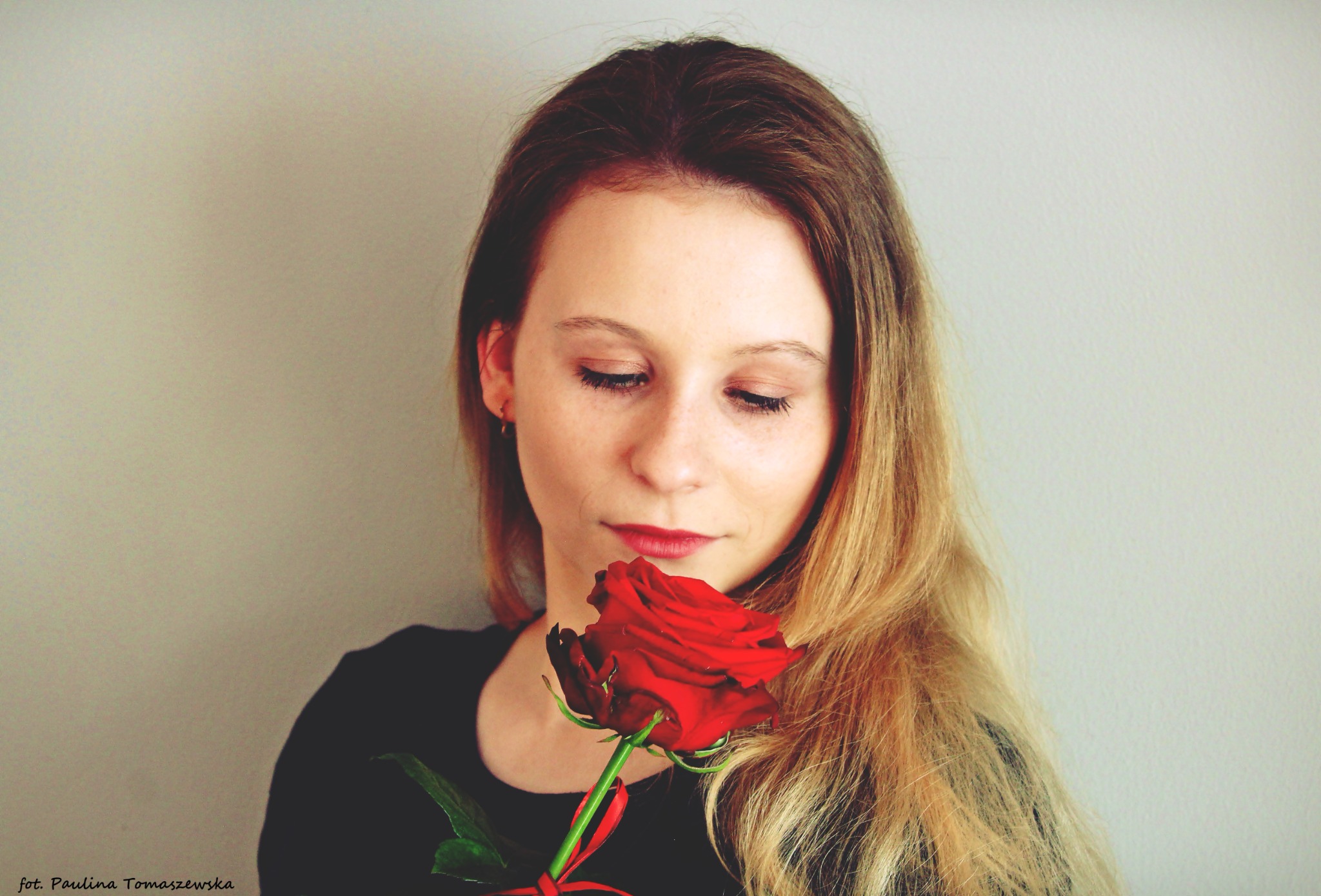 Portret młodej kobiety z długimi blond włosami, spoglądającej w dół na czerwoną różę trzymaną blisko twarzy, z delikatnym makijażem i czerwoną szminką.