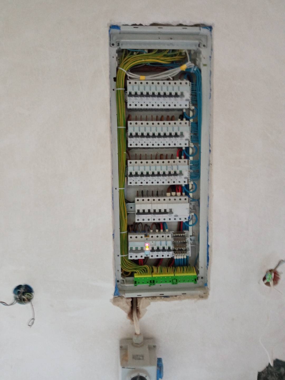 Otwarta skrzynka rozdzielcza z widocznymi zabezpieczeniami, przewodami w kolorach żółto-zielonym, niebieskim i brązowym, zamontowana w surowej ścianie z widocznymi śladami gipsu.