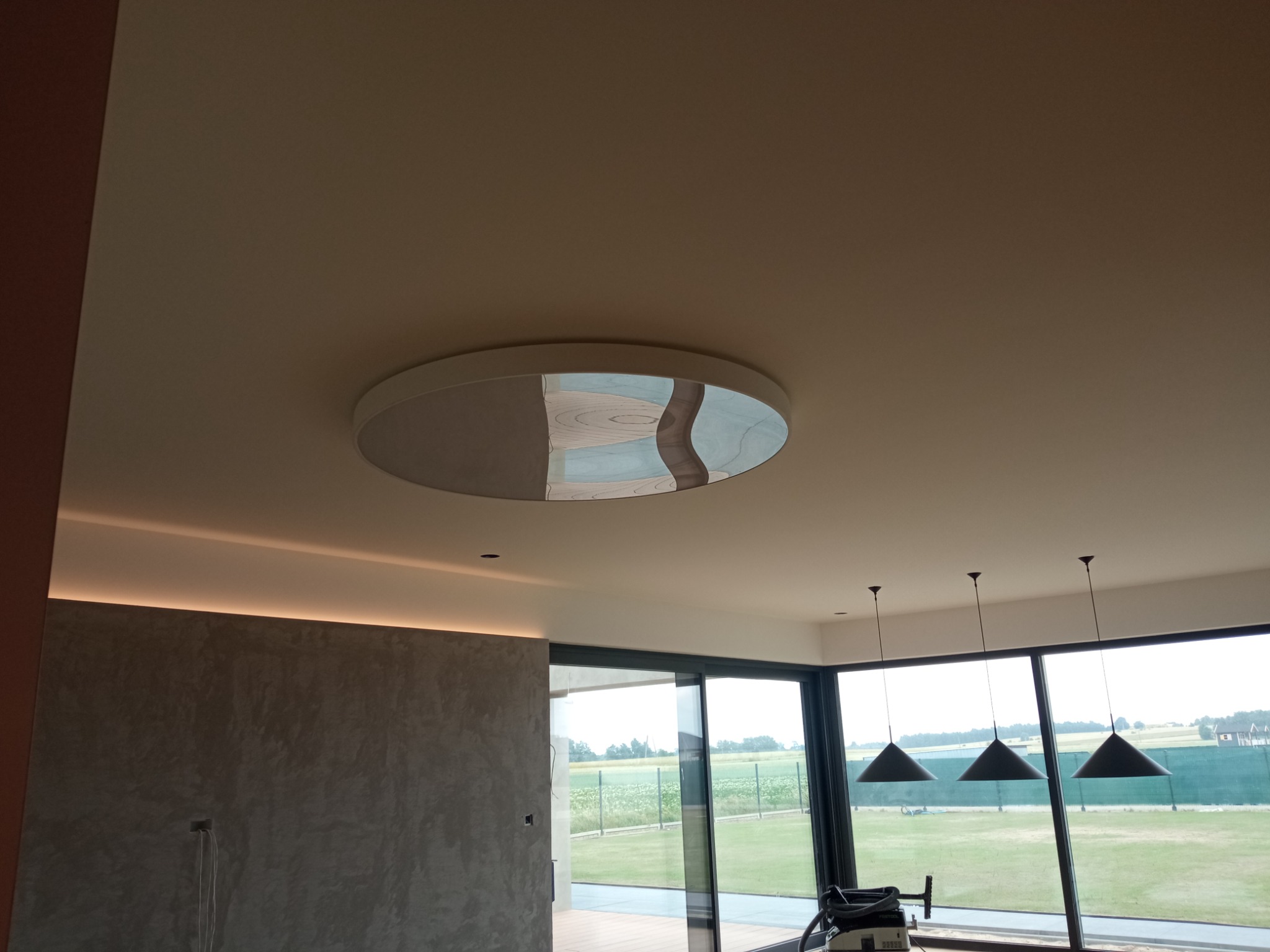 Nowoczesny salon z okrągłą lampą sufitową z efektem lustra, oświetleniem LED w listwach sufitowych i trzema czarnymi lampami wiszącymi z widokiem na zielony krajobraz za oknem.