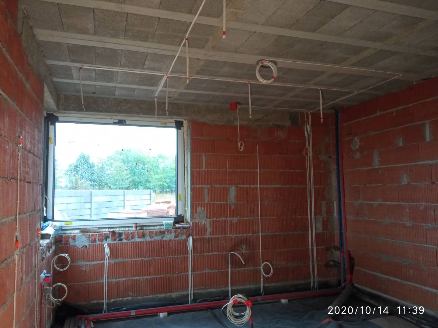 Wewnętrzne instalacje elektryczne i hydrauliczne w surowym stanie budynku z pustaków, widoczne przewody i rury, okno z widokiem na drzewa.