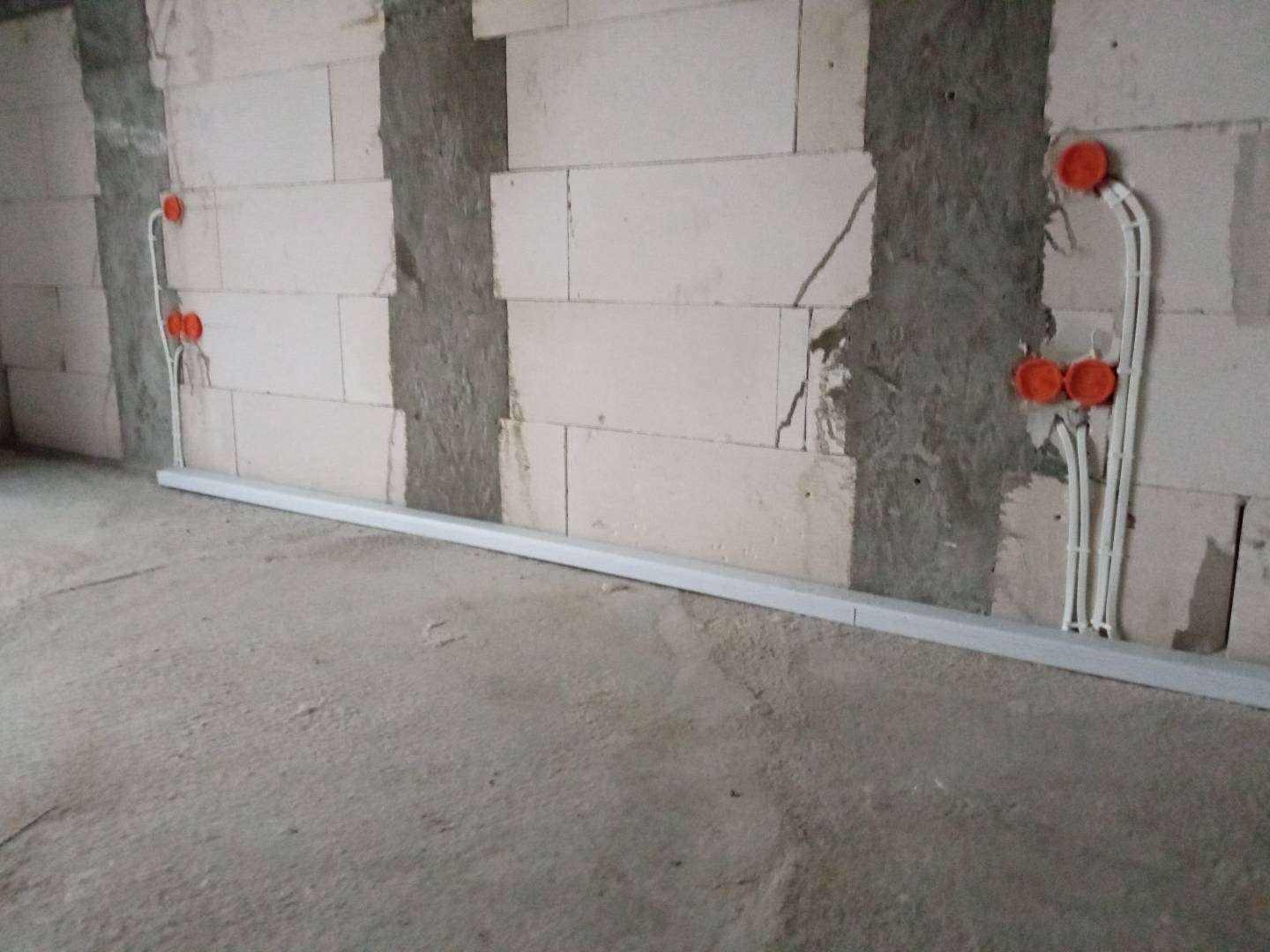 Surowy stan wnętrza: ściana z betonu komórkowego z bruzdami wypełnionymi zaprawą, widoczne pomarańczowe puszki elektryczne i białe rurki instalacyjne prowadzone wzdłuż ściany i pod sufitem.