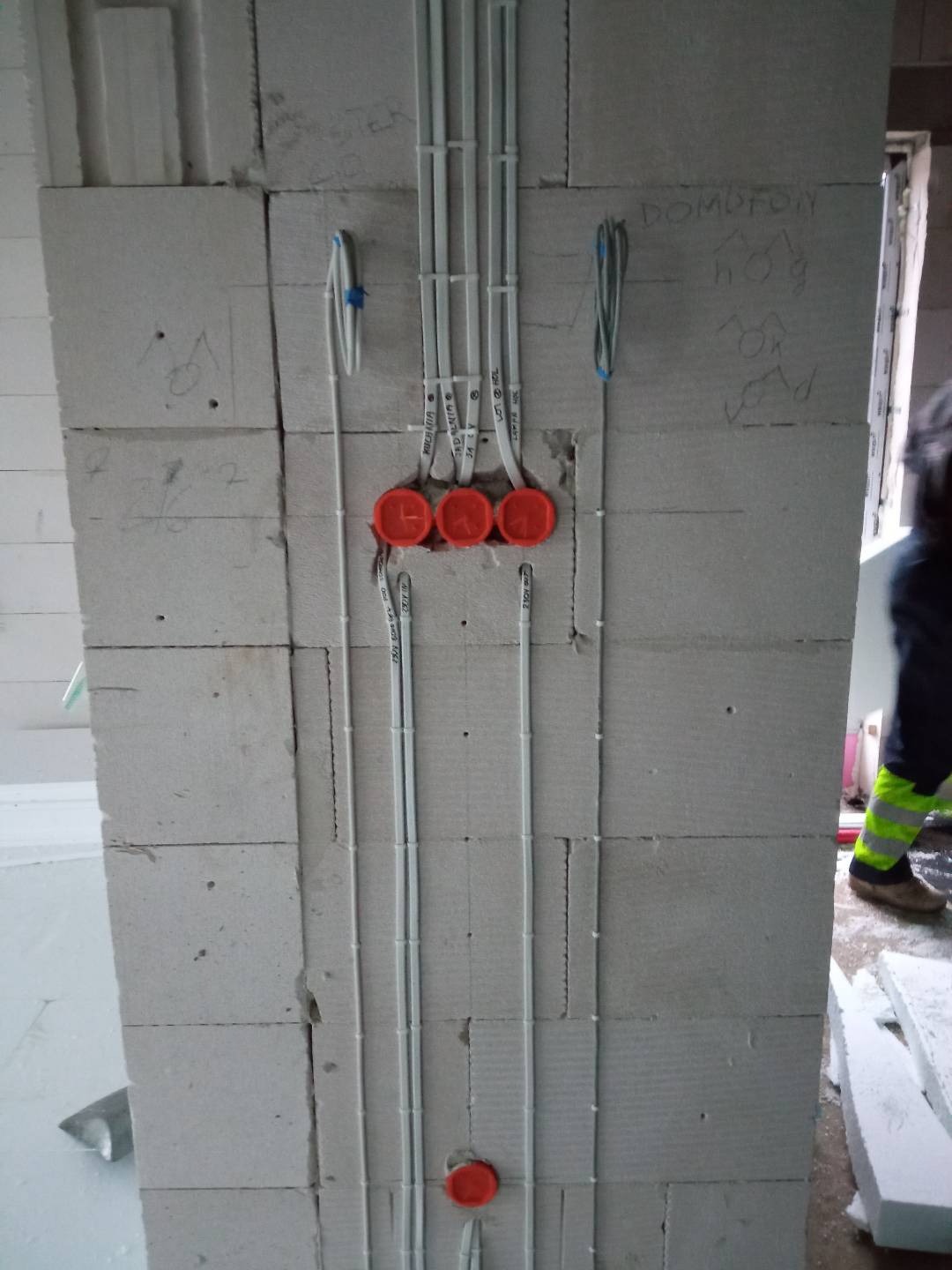 Surowa ściana z betonu komórkowego z widoczną instalacją elektryczną w peszlach i puszkach instalacyjnych, częściowo zamontowaną, z notatkami ołówkiem na ścianie.