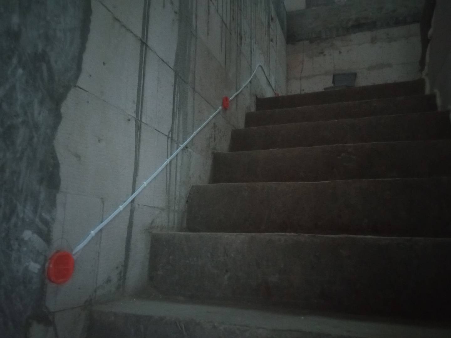 Biały kabel elektryczny przymocowany do szarej, surowej ściany klatki schodowej za pomocą czerwonych uchwytów.