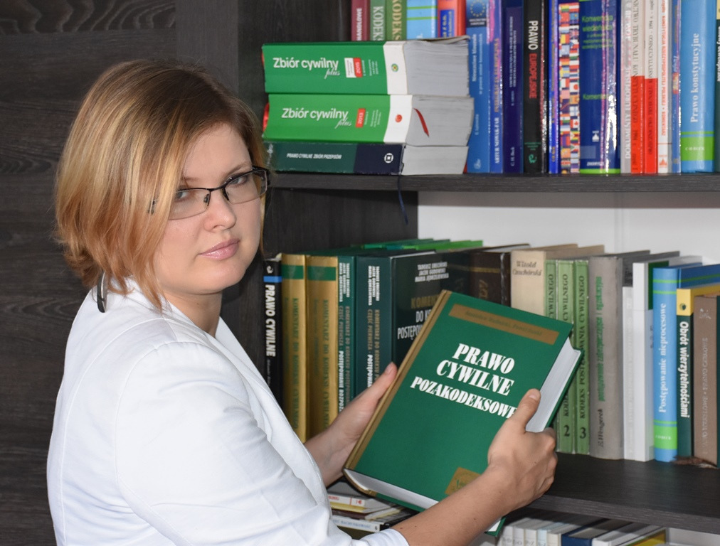 Portret kobiety w okularach, trzymającej otwartą książkę 'Prawo cywilne pozakodeksowe' na tle regału z książkami prawniczymi.