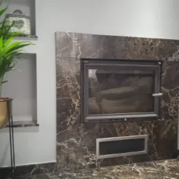 Obudowa kominka z marmuru w brązowo-białe wzory, z metalowymi drzwiczkami i wnęką wentylacyjną, obok stoi metalowy stojak na narzędzia kominkowe i roślina doniczkowa w złotym kwietniku na tle...