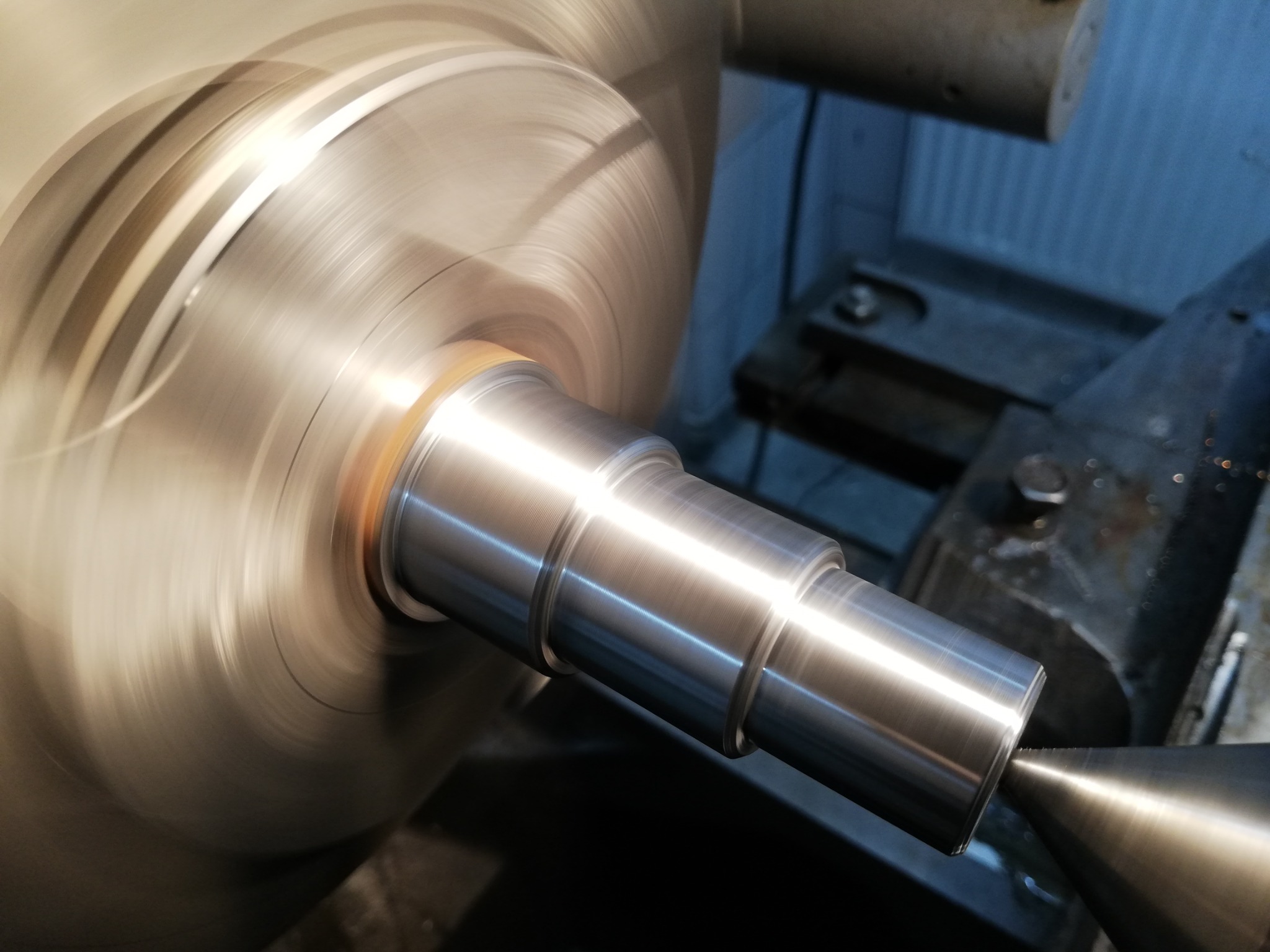 Metalowy wałek obrabiany na tokarce, widoczne wióry i narzędzie skrawające w trakcie pracy.