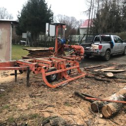 Węgrzecki Norbert - Mobilny tartak tnie kłodę drewna na deski. W tle zaparkowany pickup z załadowanym drewnem i budynek. Widoczne ślady cięcia i kora na ziemi.