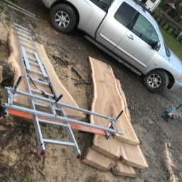 Węgrzecki Norbert - Deski z drewna leżą na ziemi obok srebrnego pickupa. Na deskach zamontowana drabina z pomarańczową prowadnicą, sugerująca wstępną obróbkę drewna.