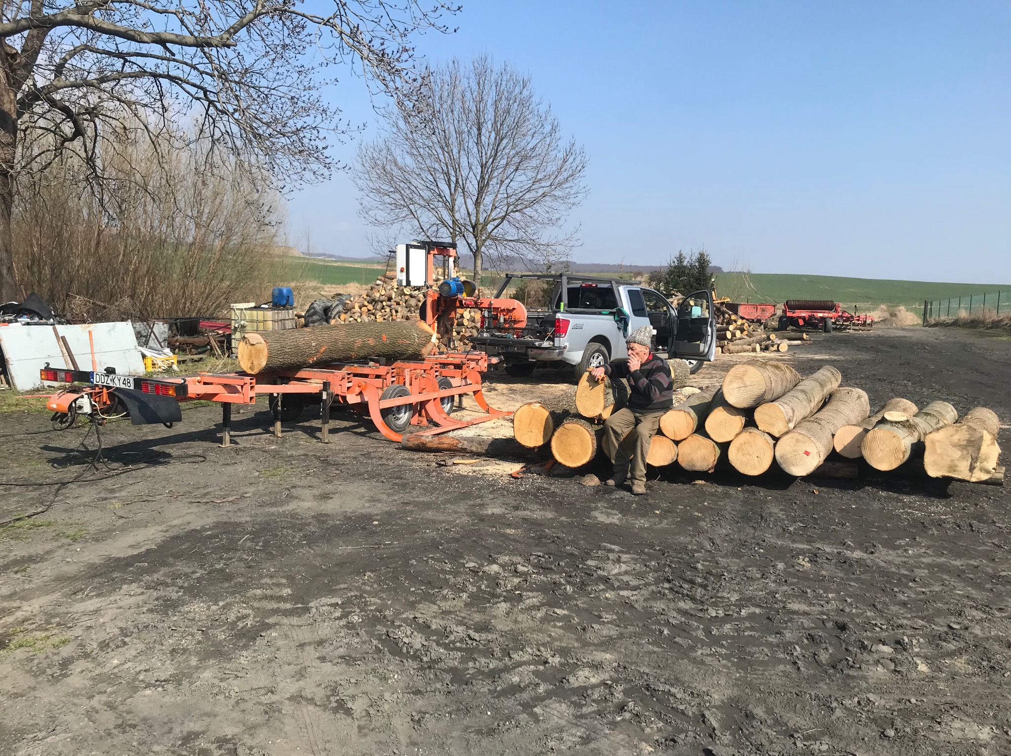 Przenośny tartak z pniem drzewa na prowadnicy, obok stos pociętych kłód i mężczyzna w czapce trzymający kawałek drewna. W tle krajobraz polny.