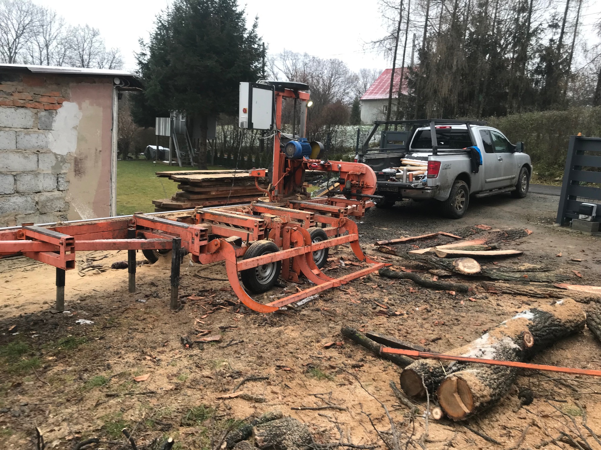 Mobilny tartak tnie kłodę drewna na deski. W tle zaparkowany pickup z załadowanym drewnem i budynek. Widoczne ślady cięcia i kora na ziemi.