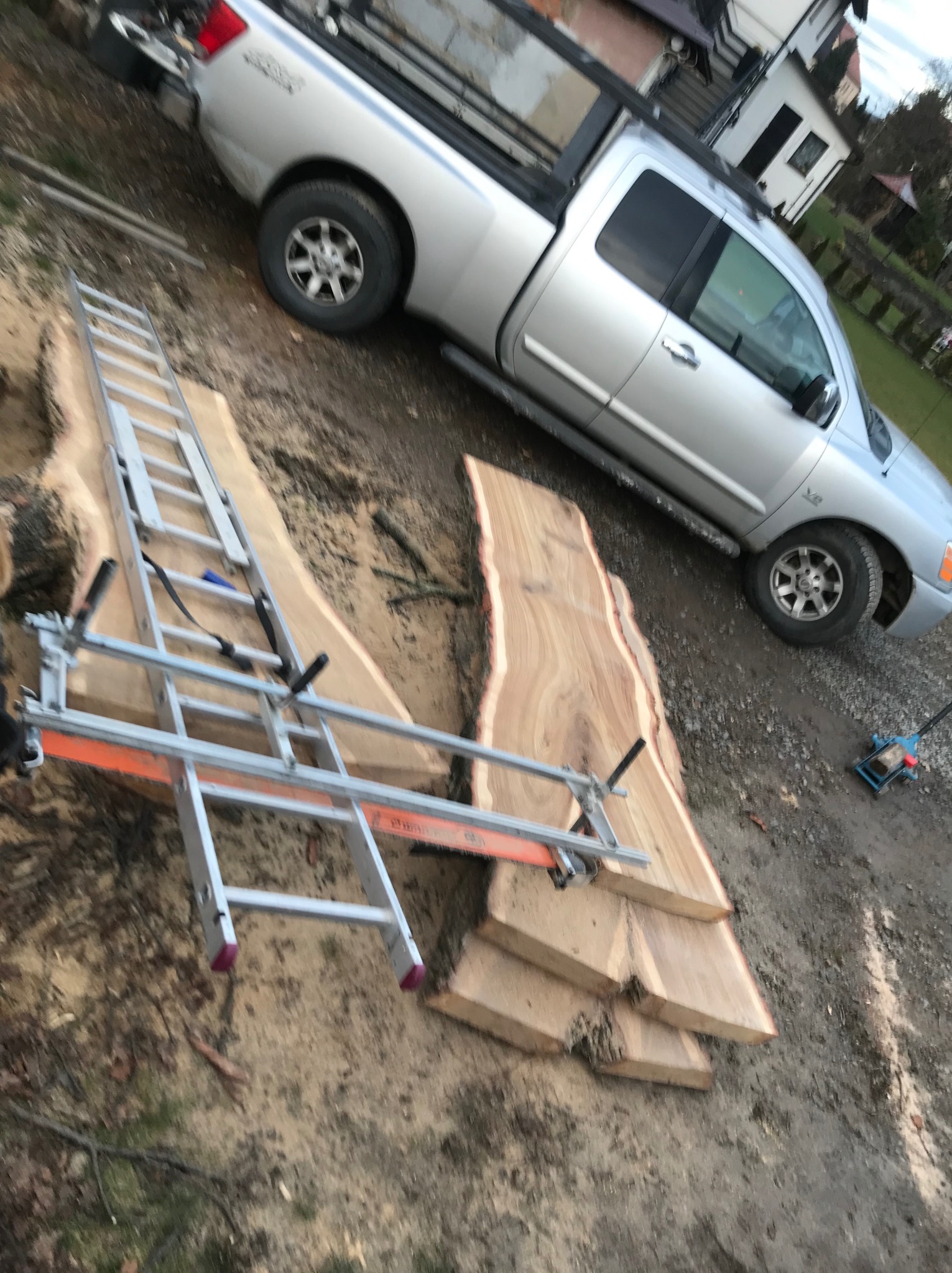 Deski z drewna leżą na ziemi obok srebrnego pickupa. Na deskach zamontowana drabina z pomarańczową prowadnicą, sugerująca wstępną obróbkę drewna.