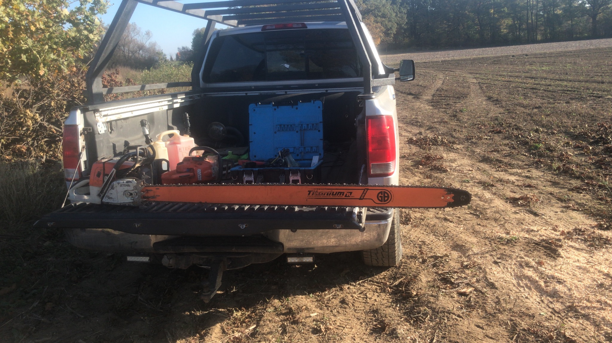 Pick-up z piłą łańcuchową, kanistrami i narzędziami na tle pola. Widok na otwartą skrzynię ładunkową z wyposażeniem do wycinki drzew.