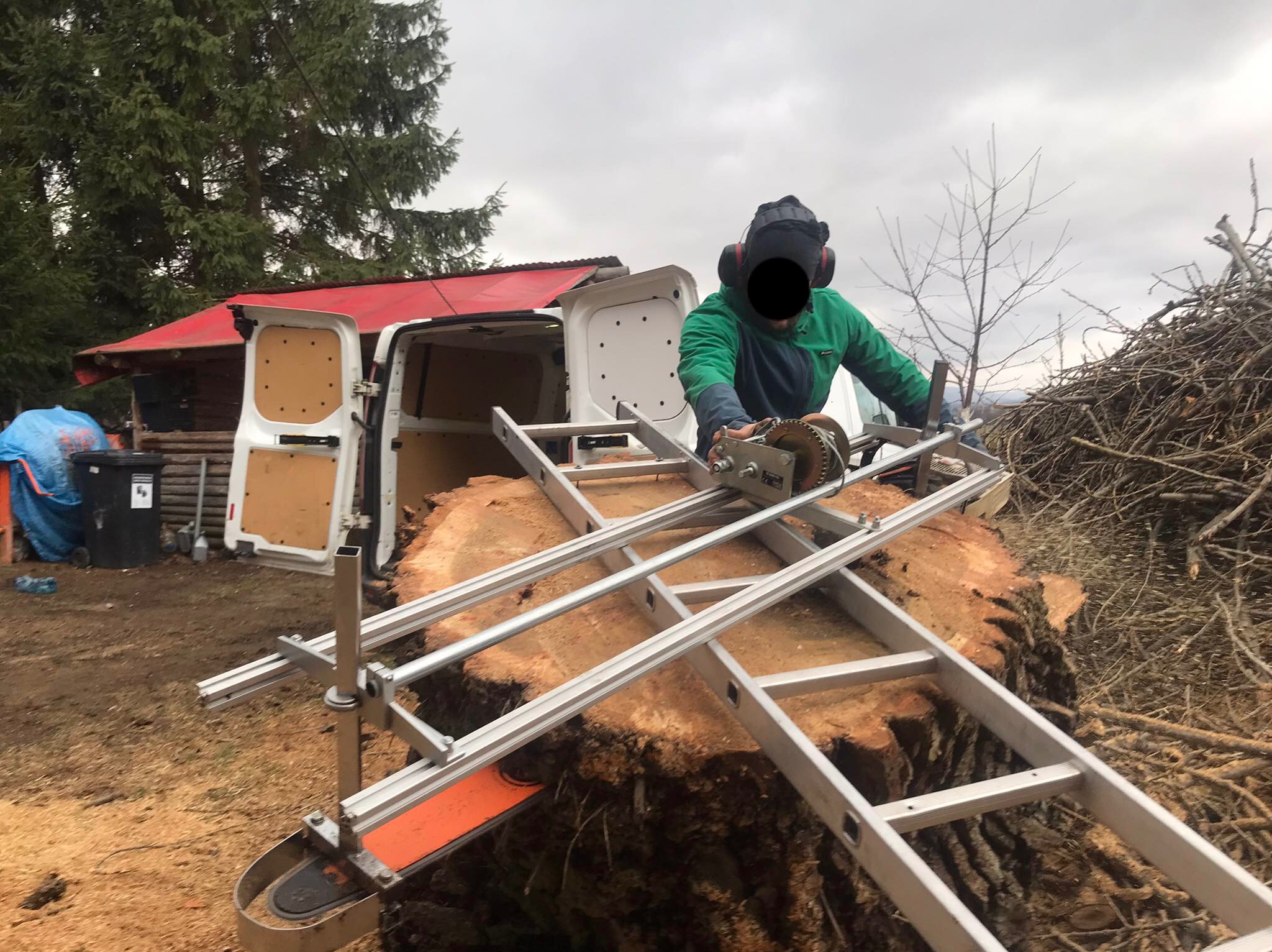 Pracownik w kasku i słuchawkach tnie kłodę piłą z prowadnicą na deski. W tle bus, budynek z czerwonym dachem i sterta gałęzi.