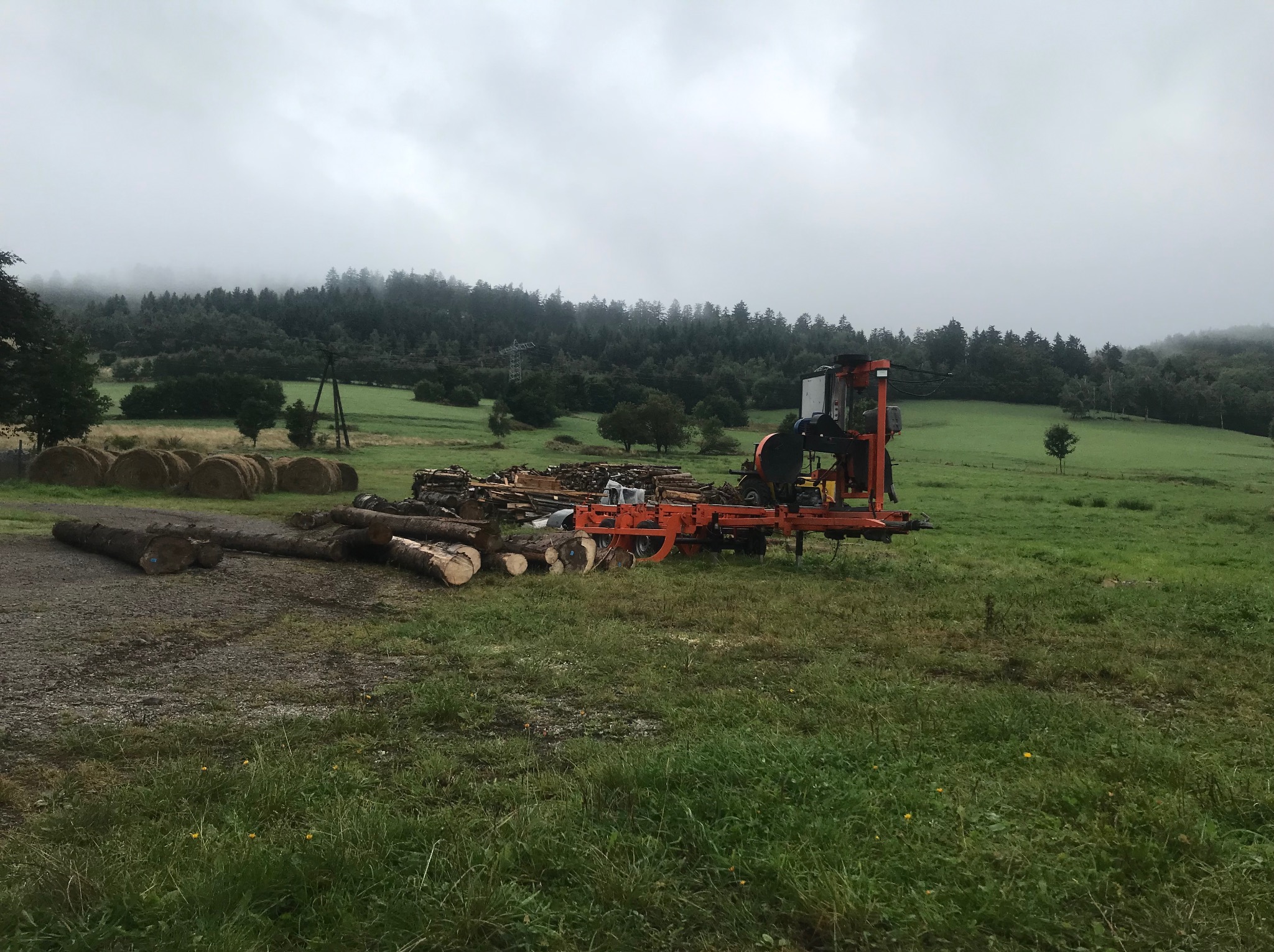Mobilny tartak w akcji na zielonej łące, w tle las i zamglone wzgórza. Stosy pociętych kłód i balotów siana obok maszyny.