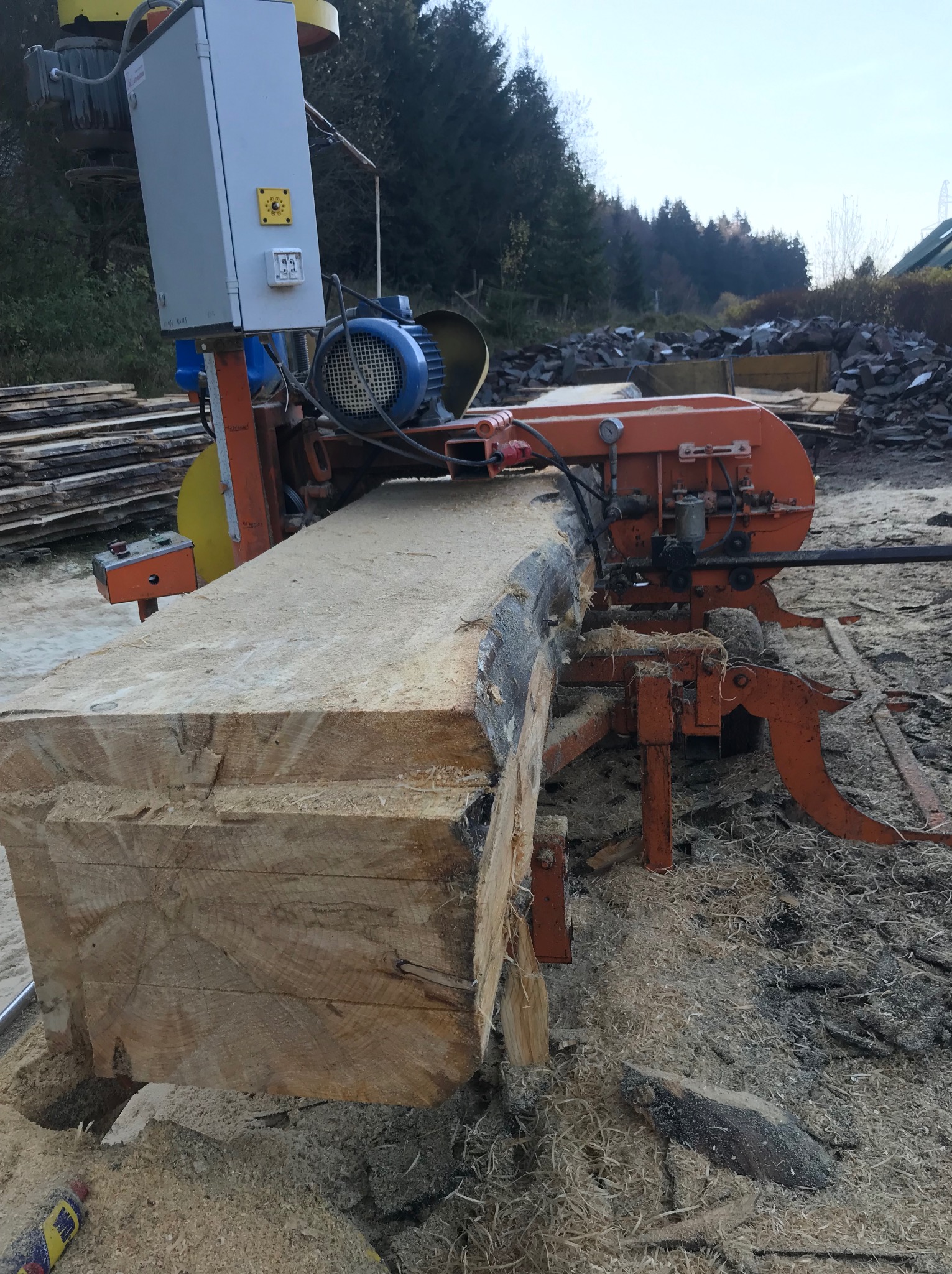 Piła taśmowa tnie gruby pień drzewa na deski. Wokół leżą wióry. W tle widać stos desek i zwałowisko kamieni.