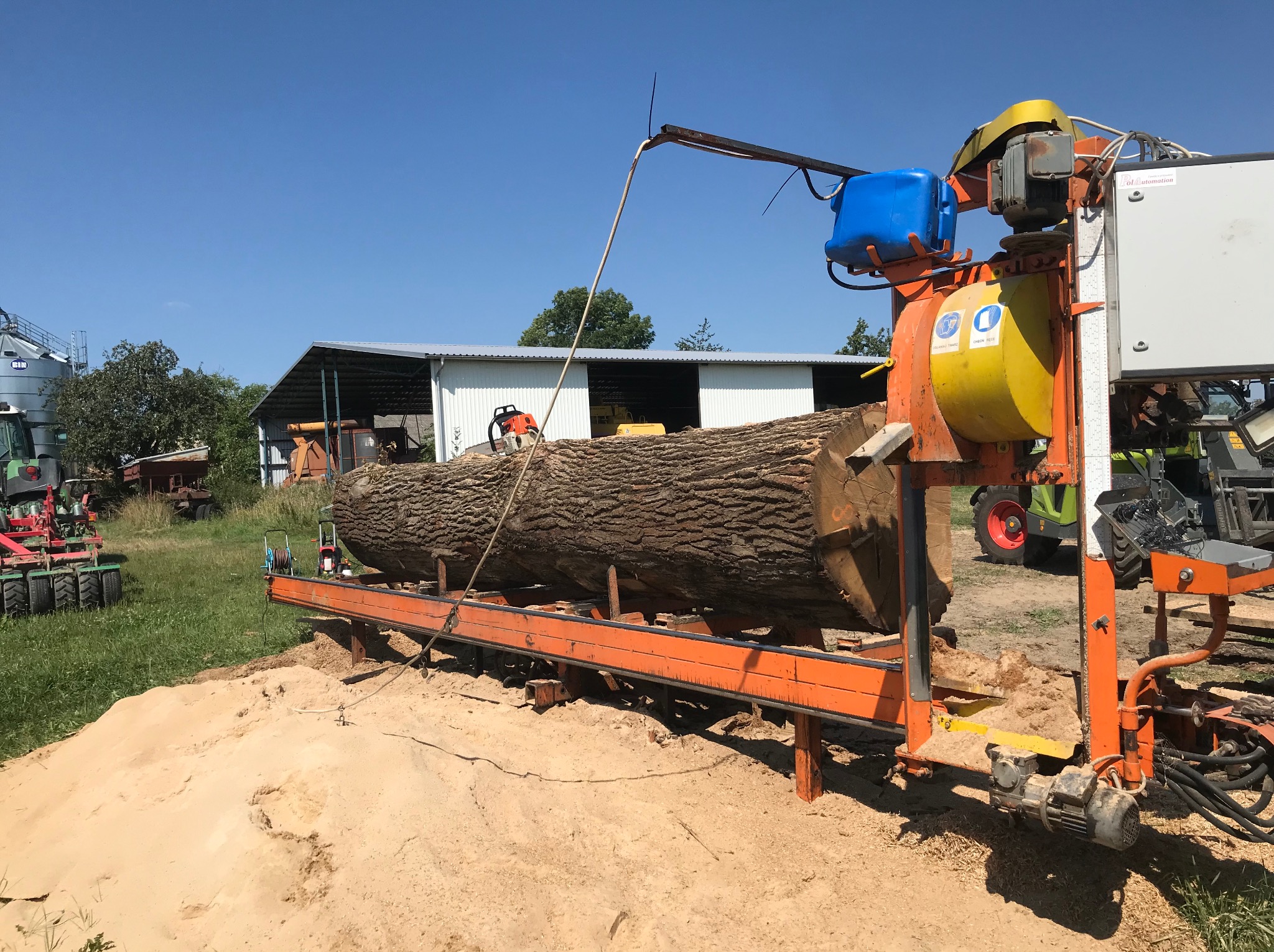 Kłoda drewna na pomarańczowej traku pionowym. W tle budynek gospodarczy, traktor i niebieskie niebo. Widoczne trociny i piła łańcuchowa na kłodzie.