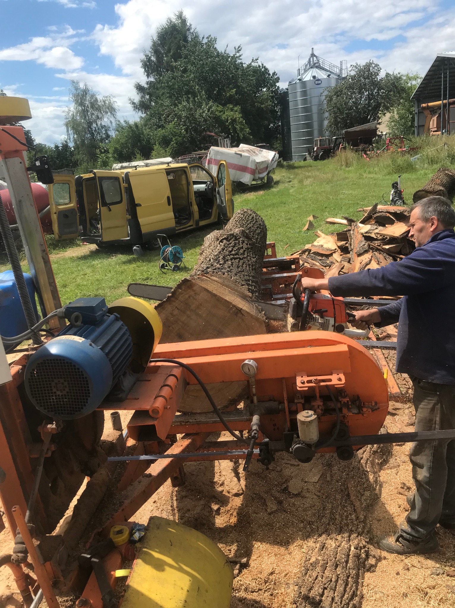 Mężczyzna obsługuje pomarańczową trakę taśmową, tnąc kłodę. W tle żółty bus i silosy zbożowe. Wokół leżą wióry.