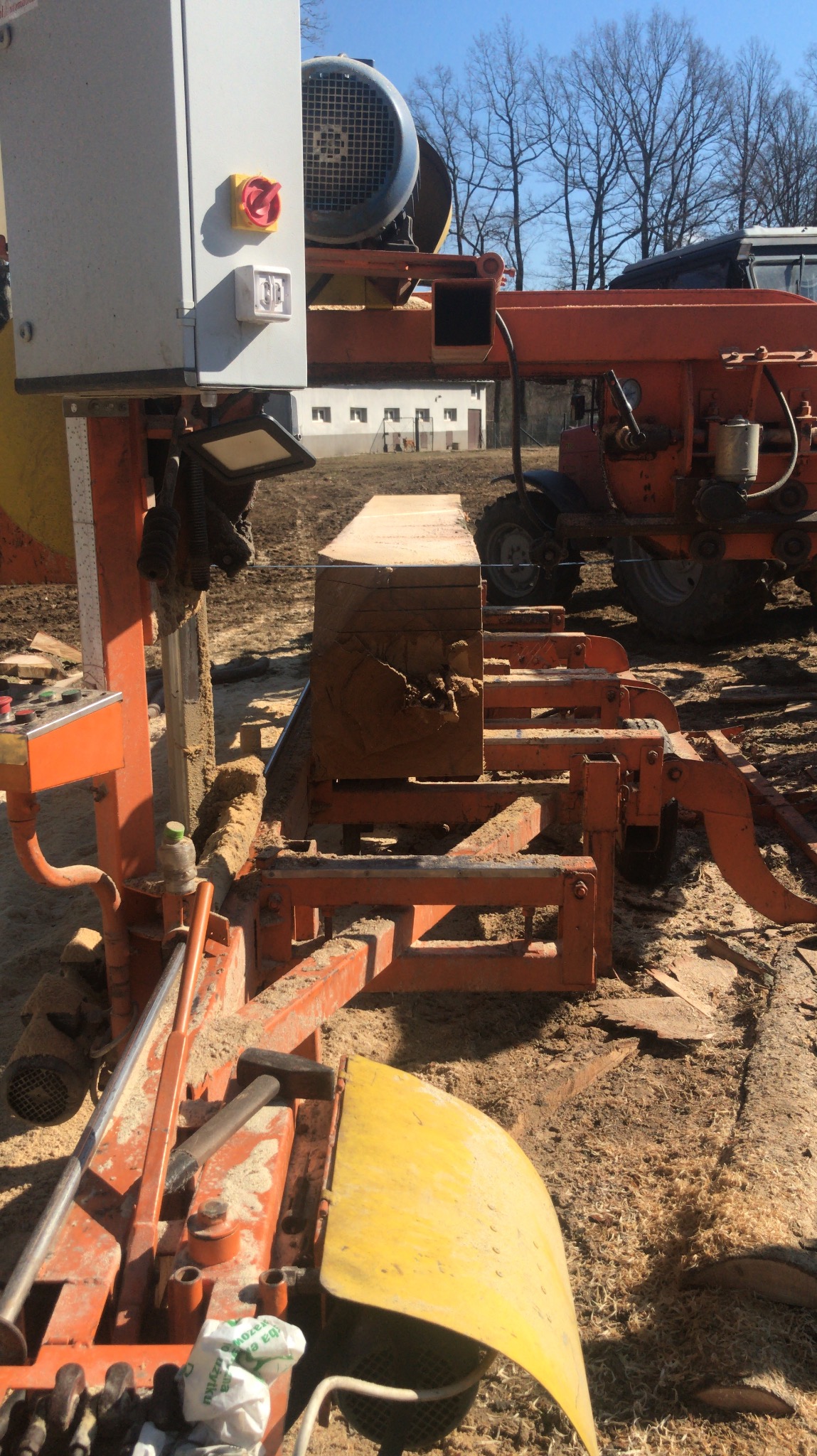 Pomarańczowa traka taśmowa tnąca drewno na deski. Widoczny blok drewna, silnik, osłona tarczy i traktor w tle. Wokół rozsypane trociny.