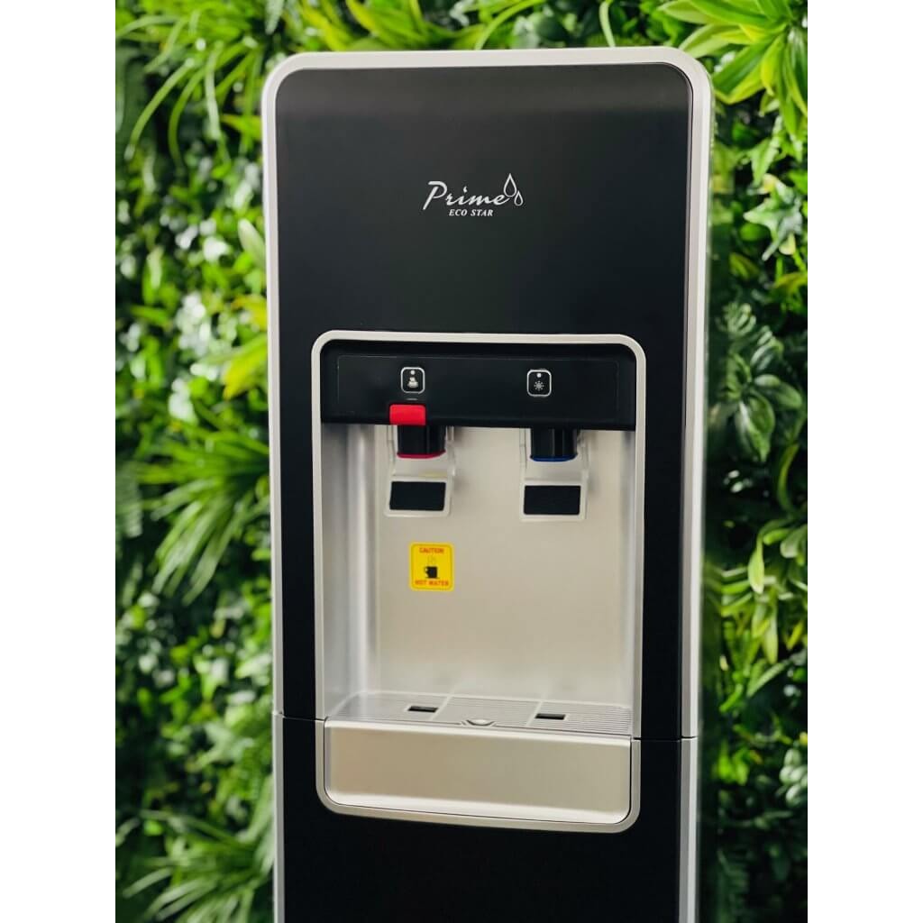 Czarny dystrybutor wody Prime Eco Star z kranikami oznaczonymi na czerwono i niebiesko, na tle ściany z zielonych roślin.