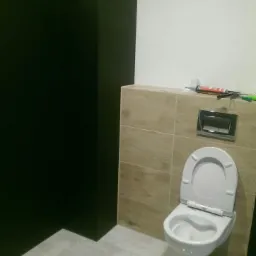 Zamontowana podwieszana toaleta w nowoczesnej łazience z płytkami imitującymi drewno i szarą podłogą, widoczne silikony i narzędzia na górze obudowy stelaża.