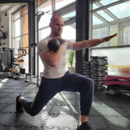 Mężczyzna w sportowym stroju wykonuje wykrok z kettlebell w siłowni, z widocznym sprzętem do ćwiczeń w tle i dużymi oknami wpadającymi naturalne światło.