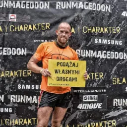 Mężczyzna po biegu przeszkodowym w Częstochowie, trzymający tabliczkę z napisem 'Podążaj własnymi drogami' na tle ściany z logotypami sponsorów.