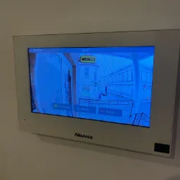 Ekran wideodomofonu Akuvox zamontowanego na ścianie, wyświetlający obraz z kamery zewnętrznej na tle ulicy z przechodniem.