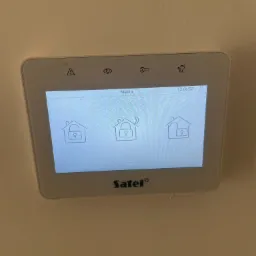 Ekran dotykowy centrali alarmowej Satel z ikonami domu i kłódki, zamontowany na beżowej ścianie.