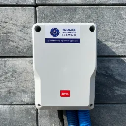 Szara skrzynka sterownicza BFT zamontowana na szarym, betonowym ogrodzeniu z widocznymi niebieskimi i czarnymi przewodami w karbowanej osłonie.