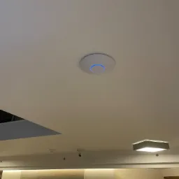 Zamontowany punkt dostępowy Wi-Fi z niebieską obwódką na suficie w biurze, widoczna kamera monitoringu i oświetlenie.