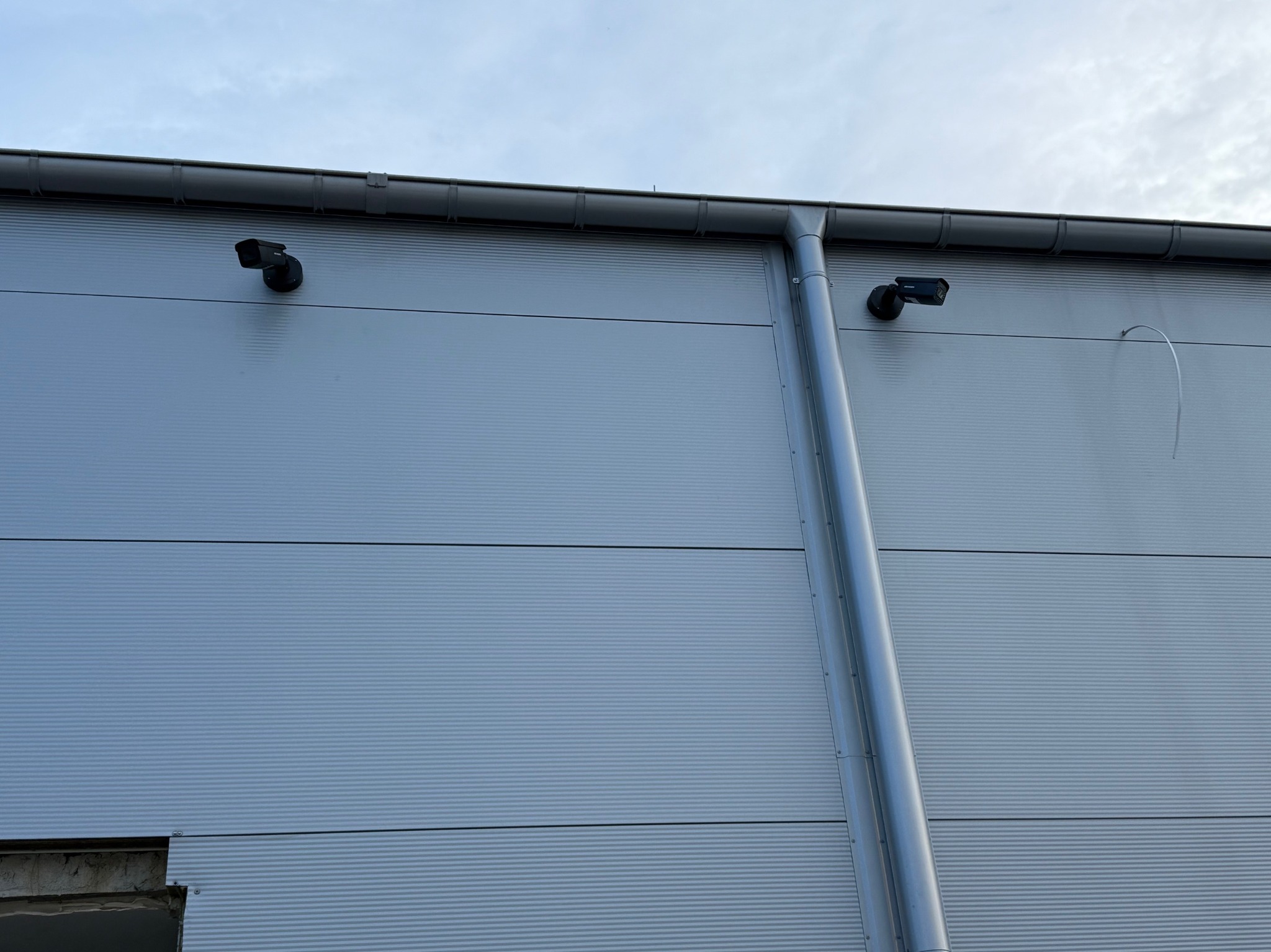 Dwie kamery Hikvision zamontowane na szarej, blaszanej ścianie budynku, obok rynny spustowej. Widoczny fragment niedokończonego otworu w ścianie.