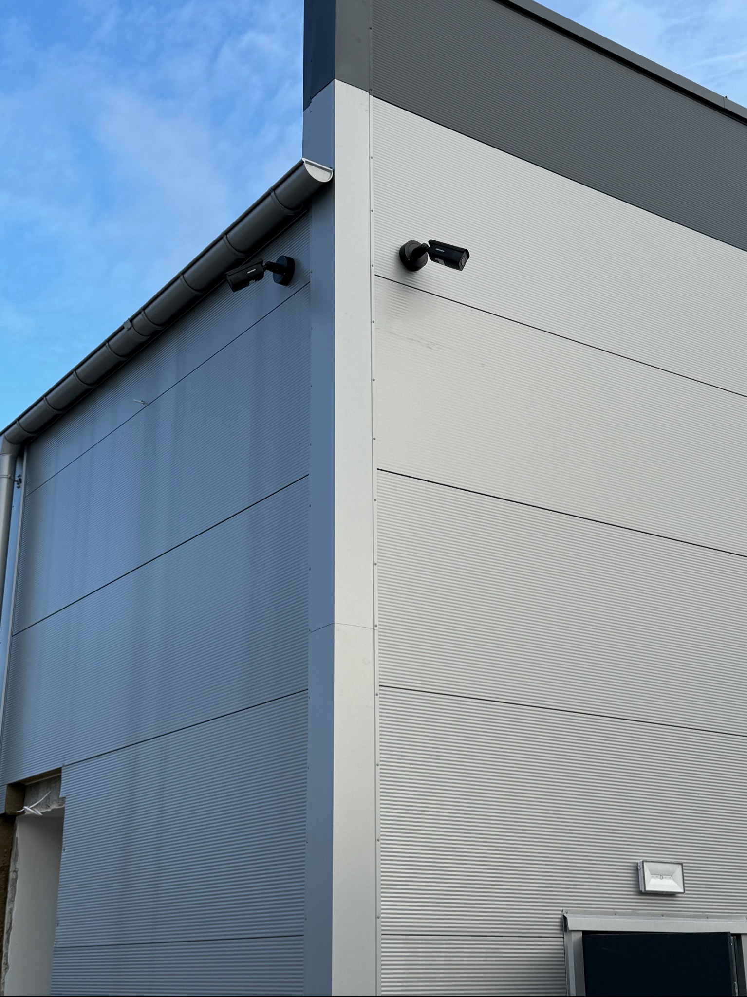 Dwie kamery Hikvision zamontowane na zewnętrznej ścianie centrum handlowego z blachy trapezowej w kolorze szarym, widok z dołu.