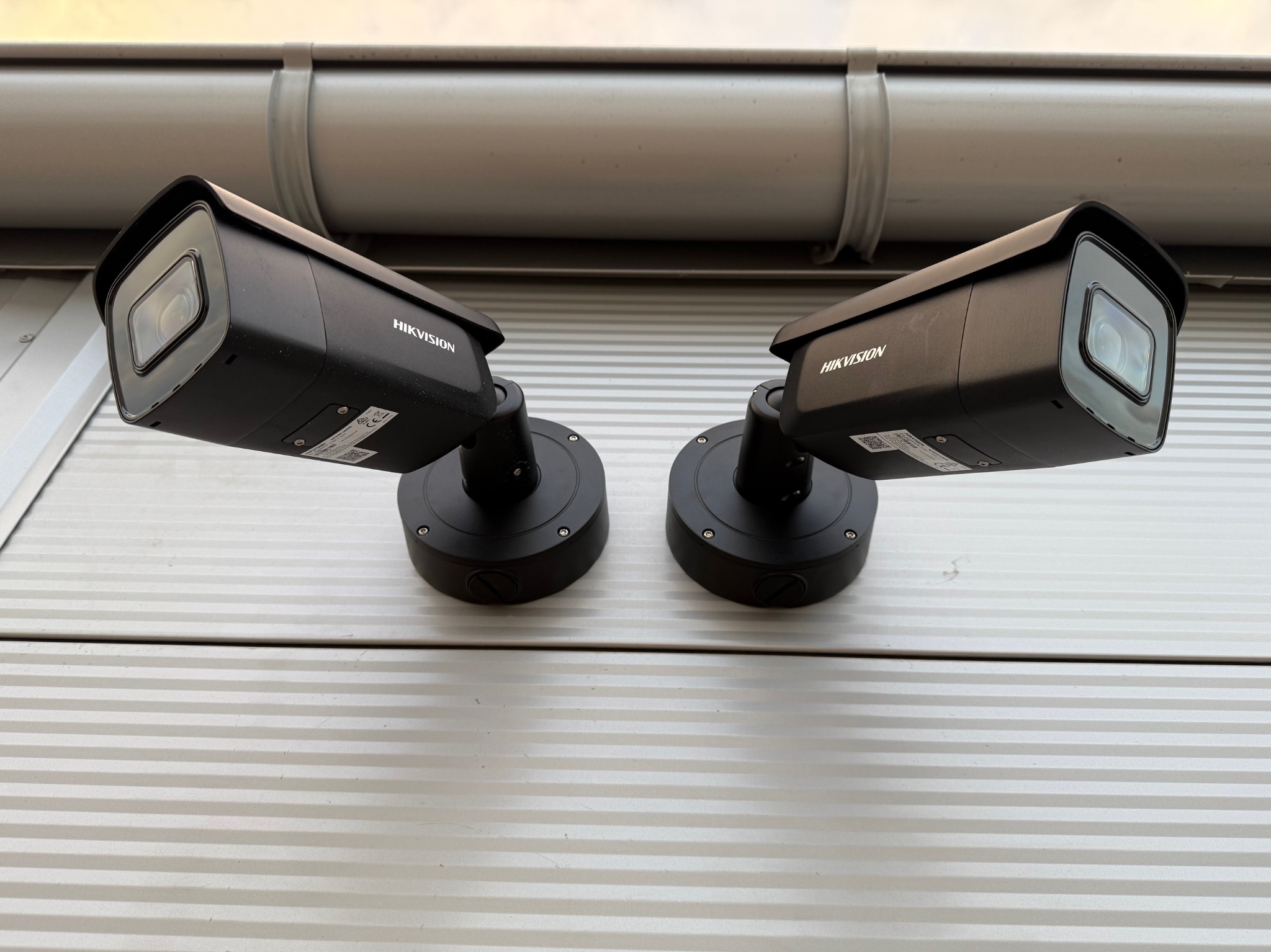 Dwa czarne kamery Hikvision zamontowane obok siebie na białej, poziomo żłobkowanej ścianie pod szarą rynną.