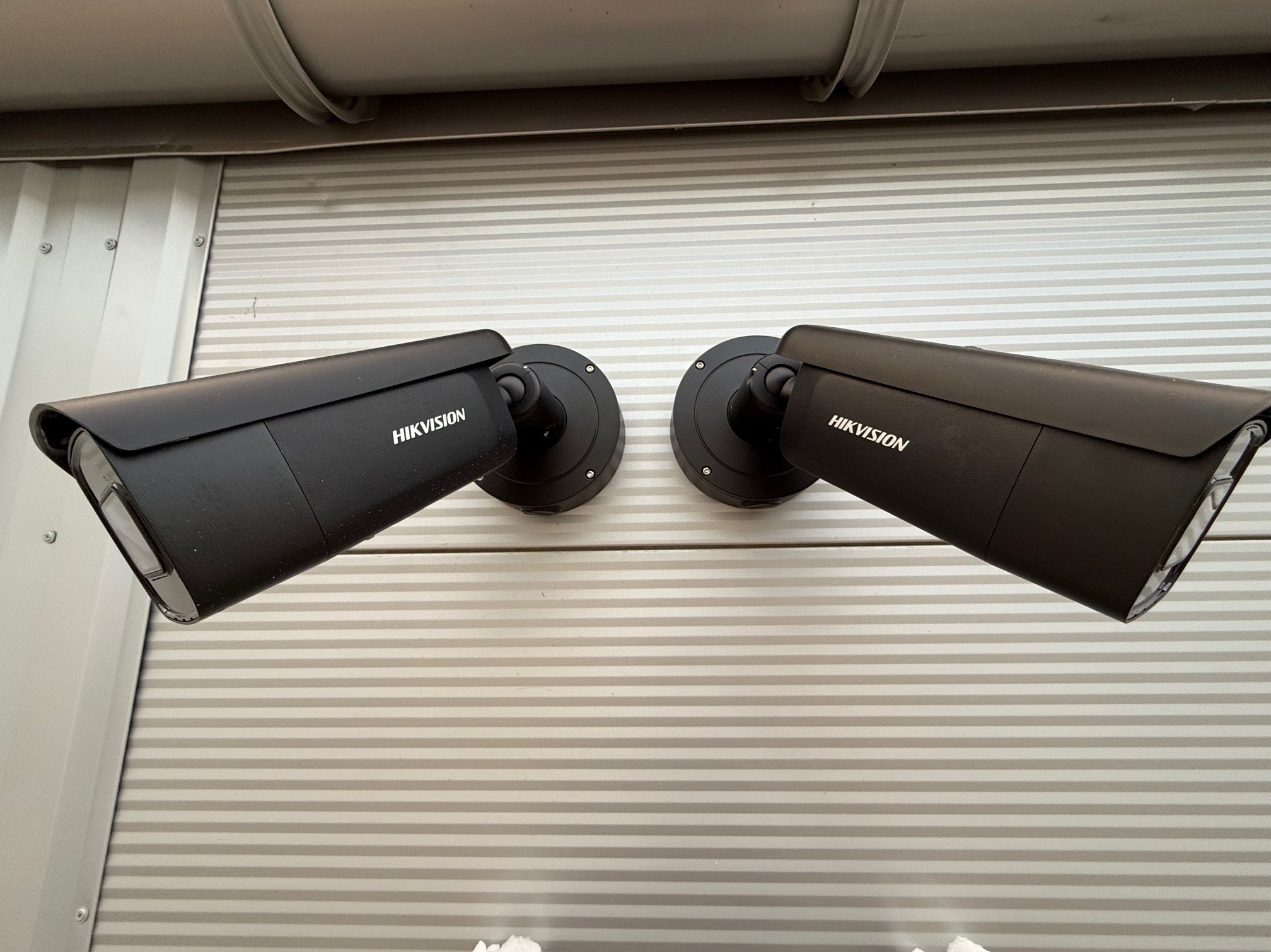 Dwie czarne kamery monitoringu Hikvision zamontowane na szarej, poziomo żebrowanej ścianie. Widoczne śruby mocujące. Kąt ujęcia lekko od dołu.