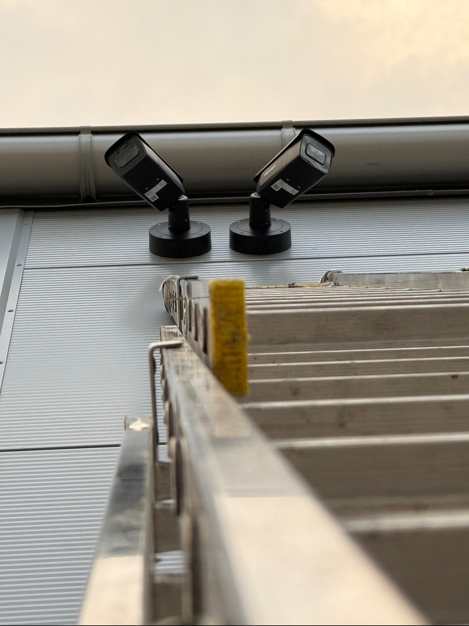 Dwie kamery Hikvision zamontowane na metalowej fasadzie budynku, widok z dołu z drabiny aluminiowej z żółtą osłoną.