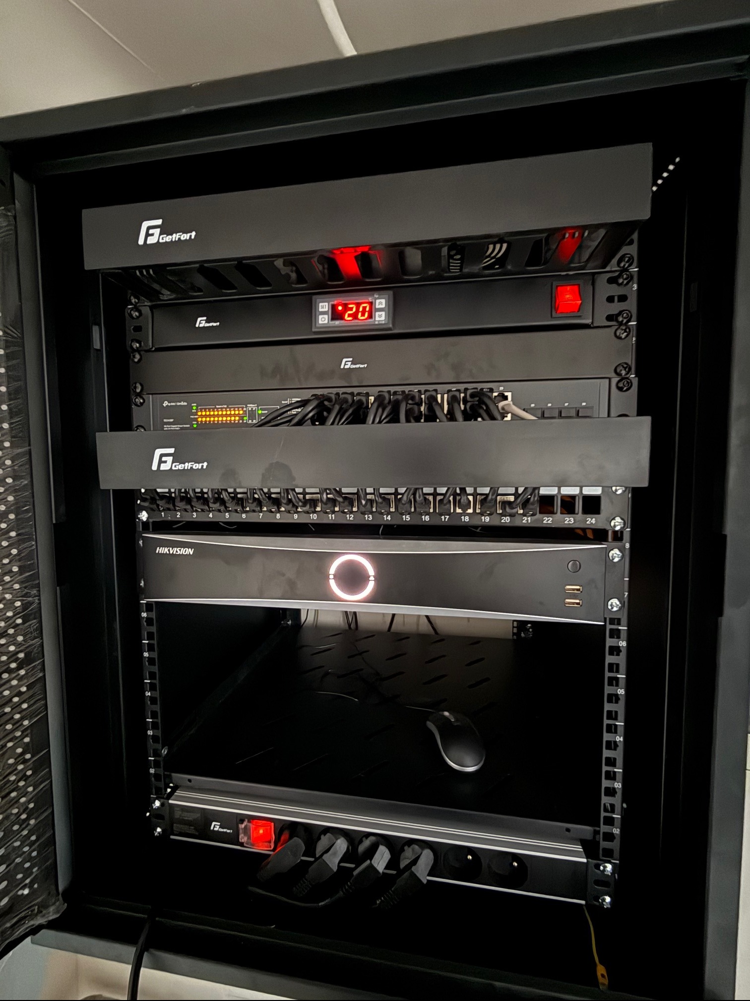 Szafa rack z zainstalowanymi urządzeniami sieciowymi i do monitoringu, w tym rejestratorem Hikvision z podświetlonym logo, routerem TP-Link z diodami oraz listwą zasilającą GetFort z włączonym...