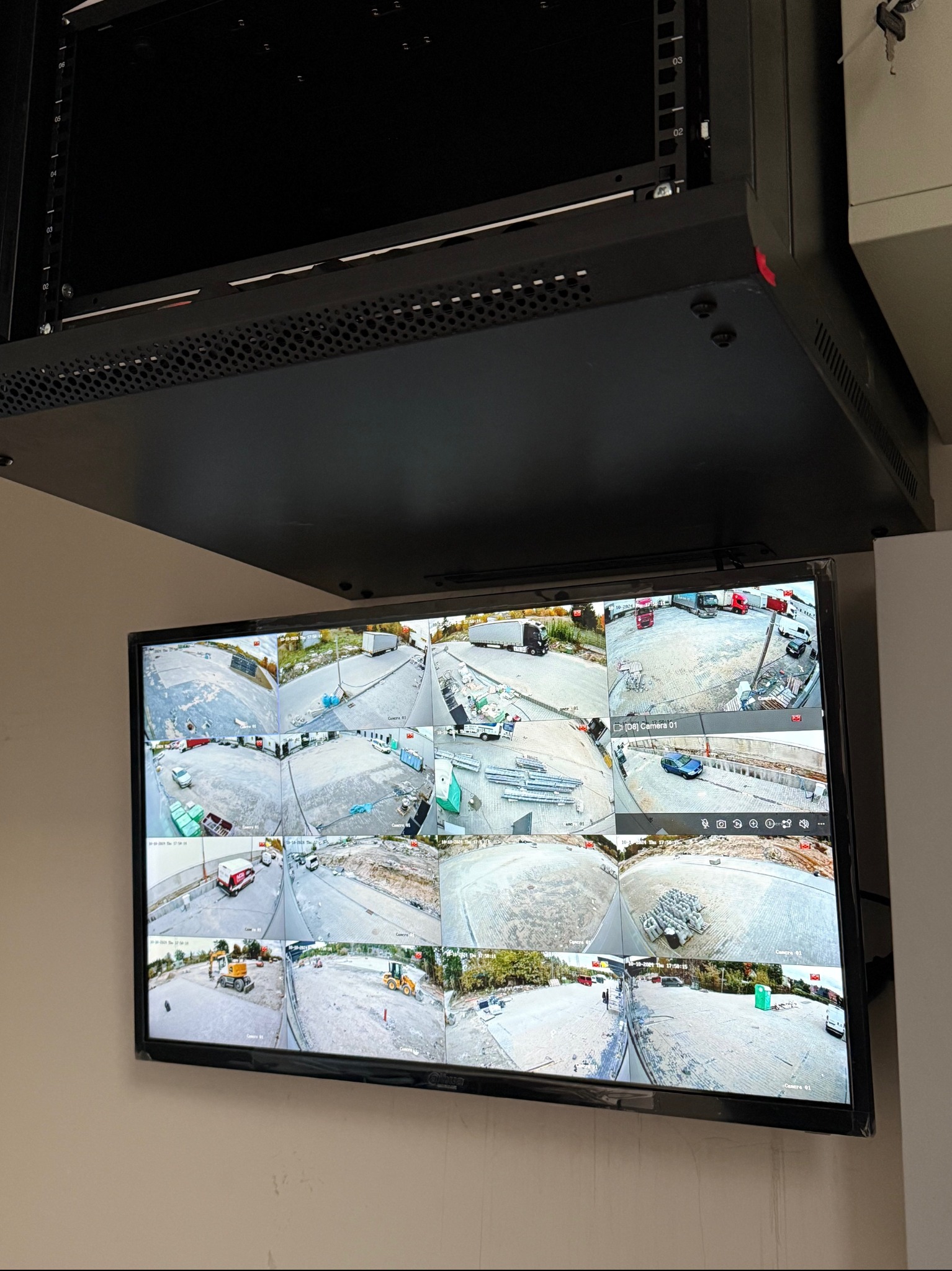 Ekran monitora z widokiem z wielu kamer przemysłowych, prezentujący plac budowy z różnymi pojazdami i materiałami, zamontowany pod szafą rack.