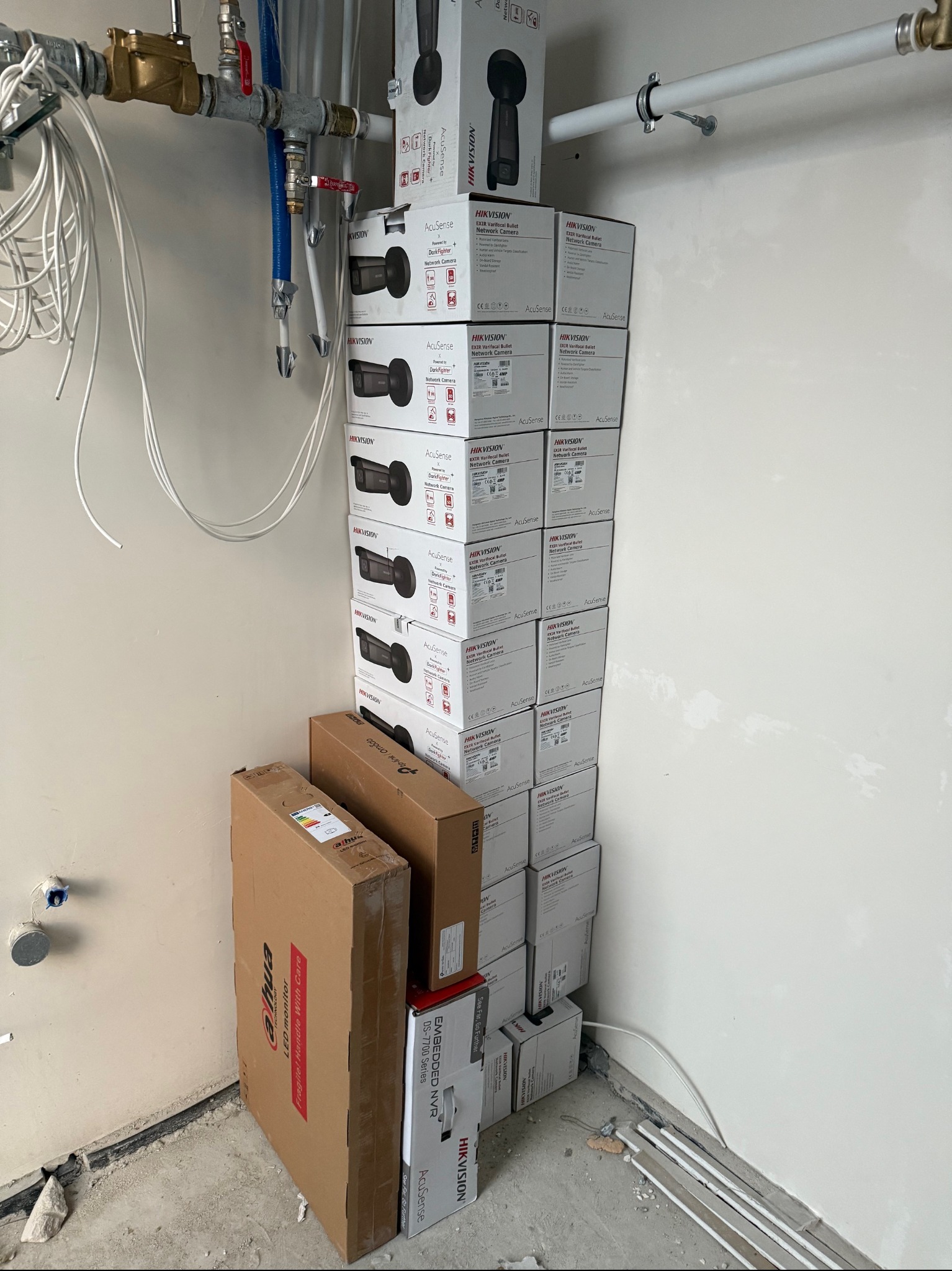 Stos pudełek z kamerami Hikvision i rejestratorami Dahua na podłodze w surowym pomieszczeniu z instalacjami hydraulicznymi na ścianie.