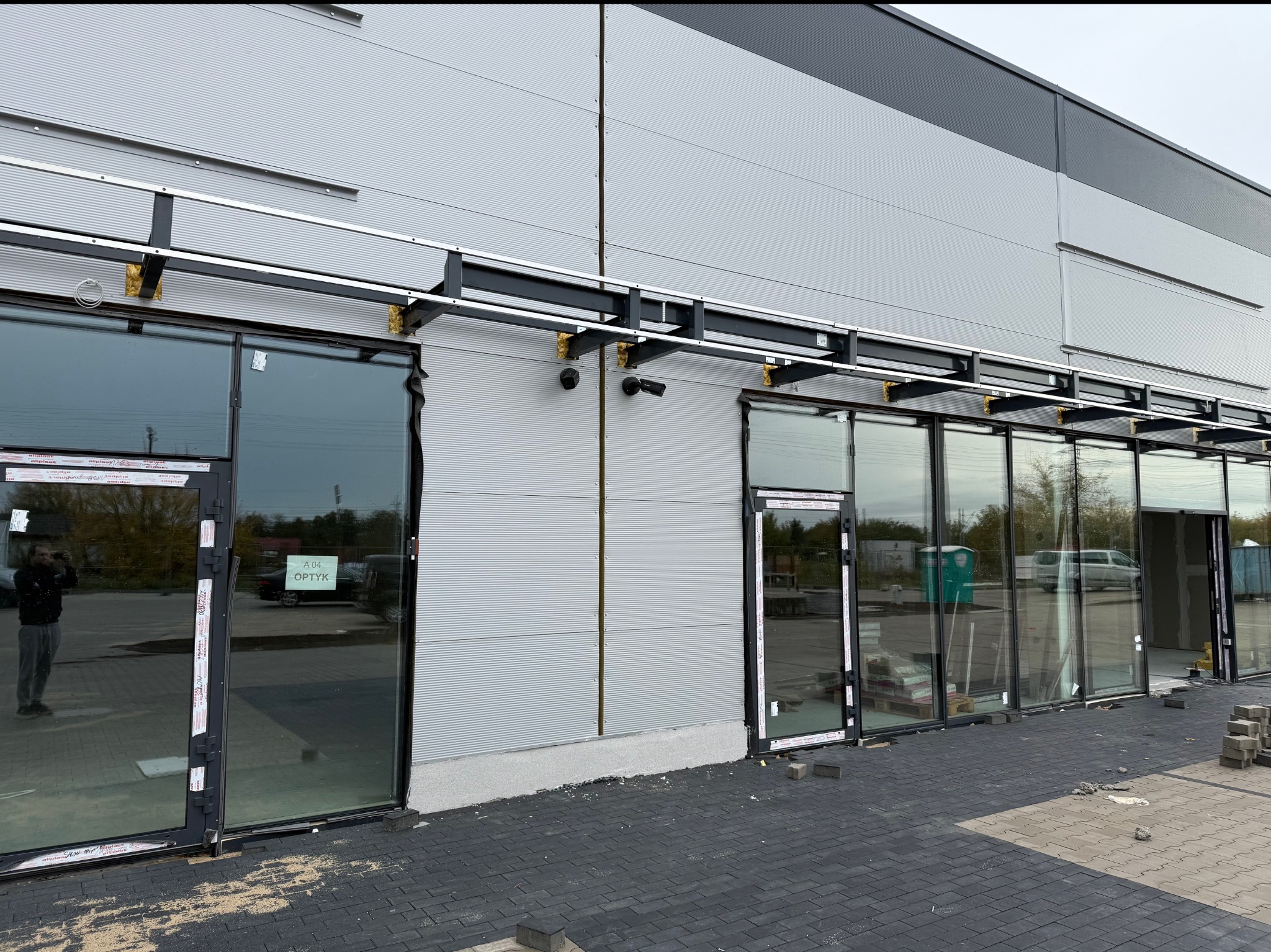 Nowoczesny budynek handlowy w trakcie wykończenia z zamontowanymi oknami i przygotowaną konstrukcją pod system kamer, widoczne kamery na elewacji, odblask fotografa w szybie.