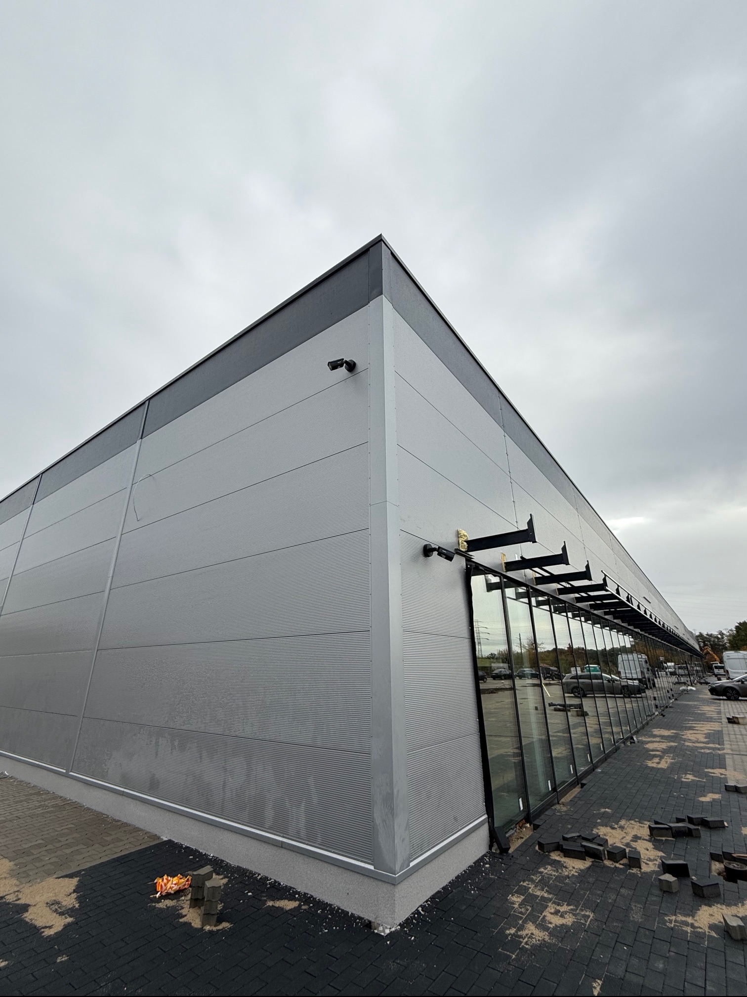 Zewnętrzny widok na szarą halę centrum handlowego z zamontowanymi kamerami i szklanymi witrynami, na chodniku porozrzucane kostki brukowe.