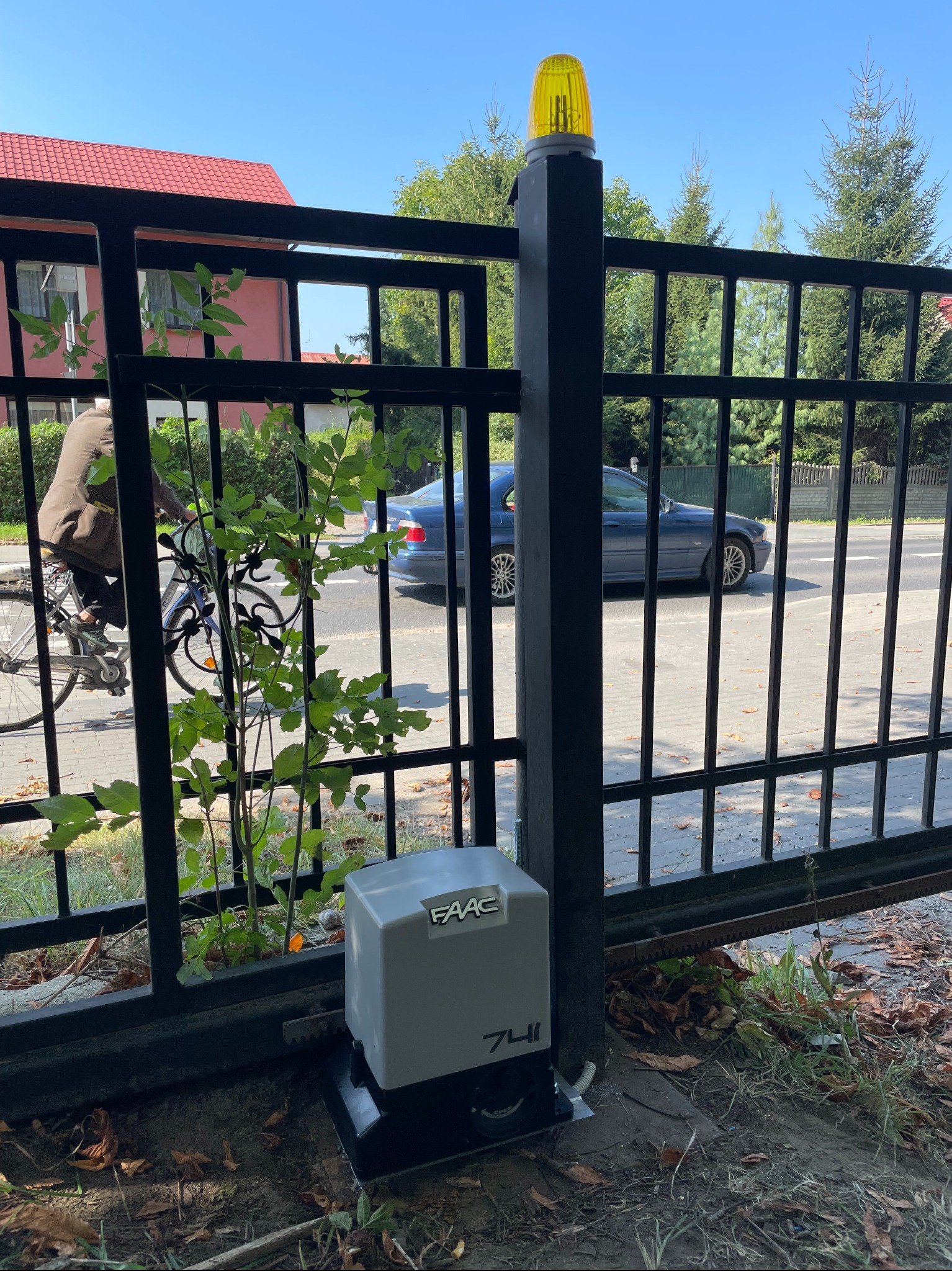 Szary napęd bramy marki FAAC 741 zamontowany przy czarnej, metalowej bramie wjazdowej, z widoczną lampą ostrzegawczą, na tle ulicy z przejeżdżającym rowerzystą i samochodem osobowym.