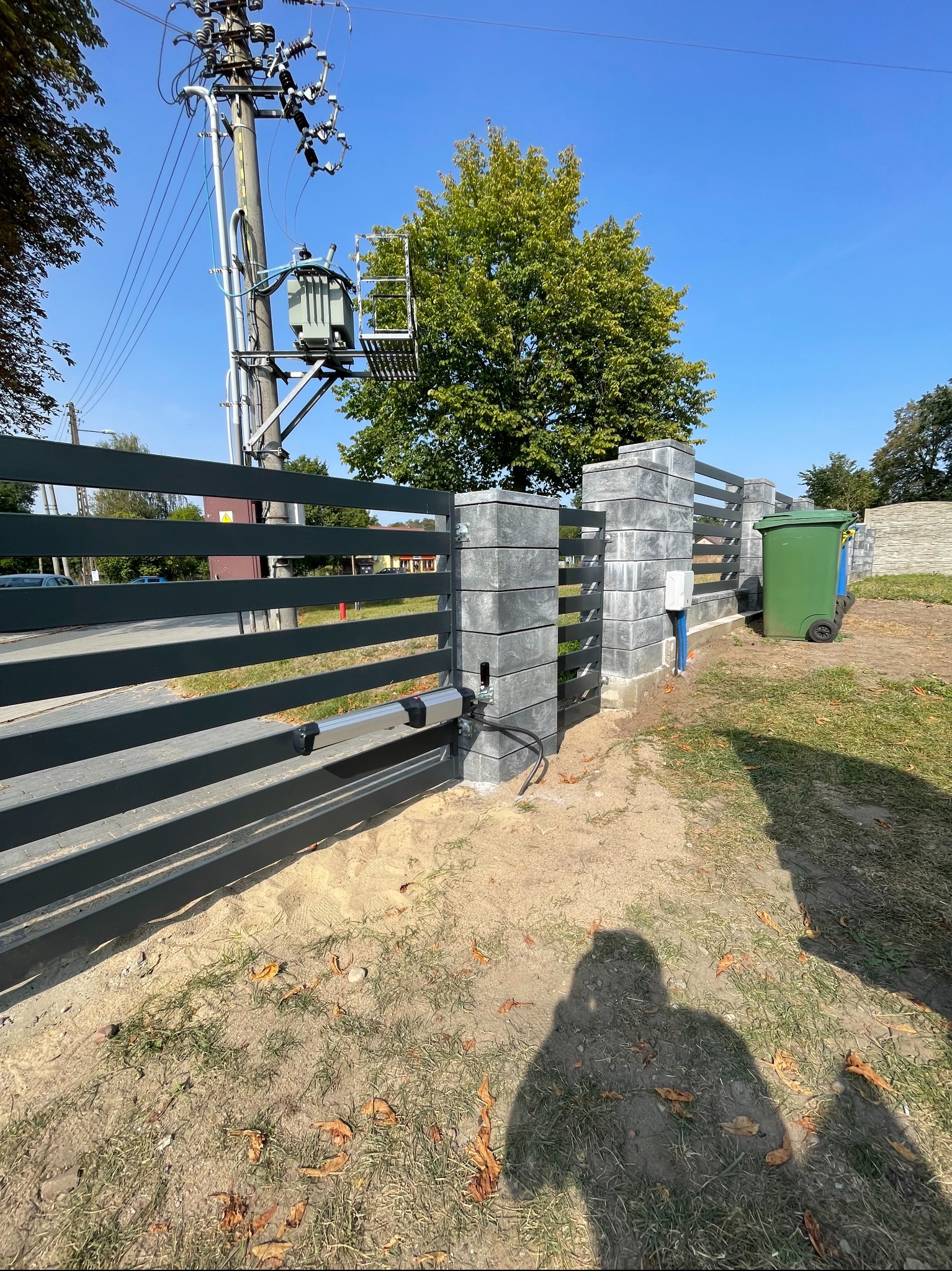 Szara, przesuwna brama wjazdowa z automatycznym napędem, zamontowana na słupkach z szarej kostki, widoczna skrzynka sterująca i kable, na tle zieleni i błękitnego nieba.