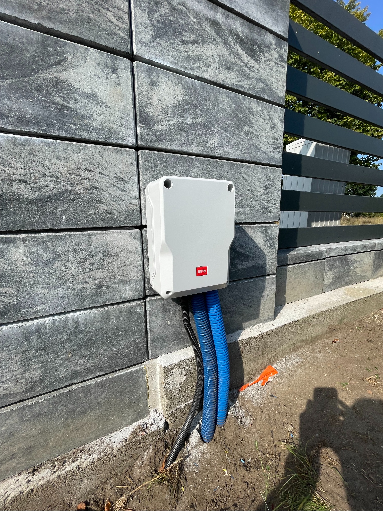 Szara skrzynka sterownicza BFT z niebieskimi i czarnymi rurami karbowanymi, zamontowana na słupku bramowym z szarej kostki, widoczny fragment ogrodzenia z poziomych profili.