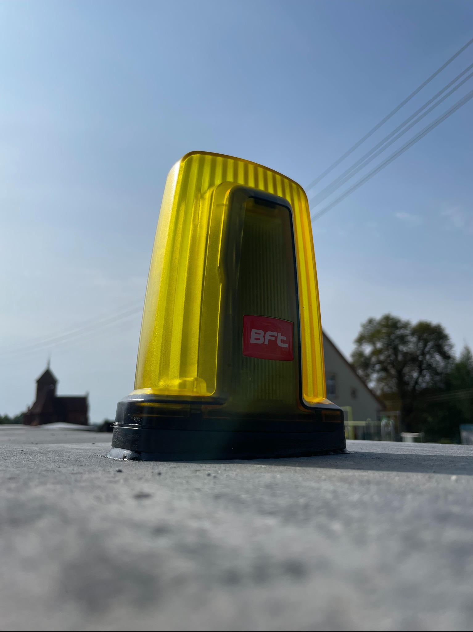 Żółta lampa ostrzegawcza BFT zamontowana na szarym podłożu w Sławnie, w tle widoczny zarys kościoła i drzewa.