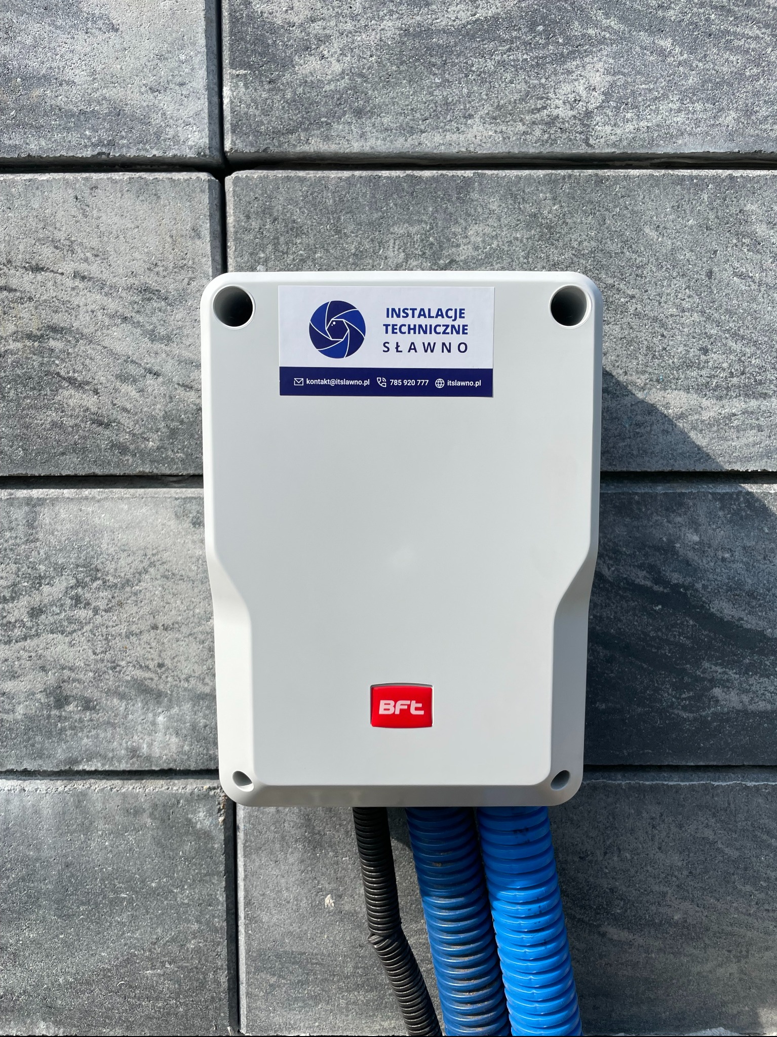Szara skrzynka sterownicza BFT zamontowana na szarym, betonowym ogrodzeniu z widocznymi niebieskimi i czarnymi przewodami w karbowanej osłonie.