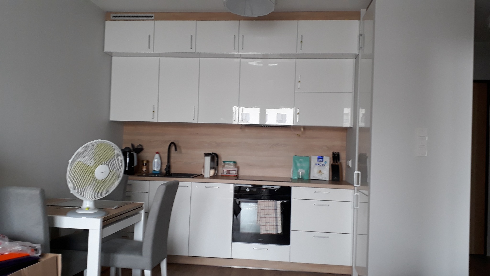 Jasna kuchnia z białymi szafkami na wymiar, brązowym panelem nad blatem i widocznym fragmentem stołu z wentylatorem.