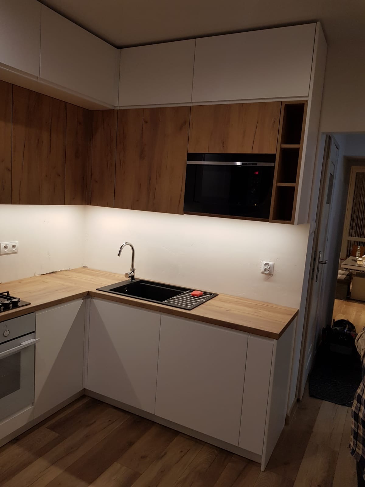 Nowoczesna kuchnia z białymi szafkami dolnymi i drewnopodobnymi górnymi, czarny zlew z ociekaczem i czerwonym zmywakiem, oświetlenie LED pod szafkami, widoczna zabudowa piekarnika.