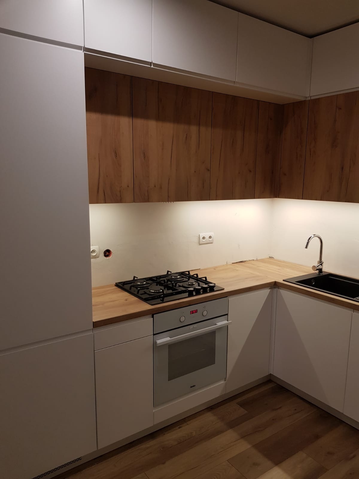 Nowoczesna kuchnia z białymi szafkami i drewnopodobnymi frontami górnych szafek, czarny zlew i płyta gazowa, oświetlenie podszafkowe, widoczne gniazdka elektryczne.